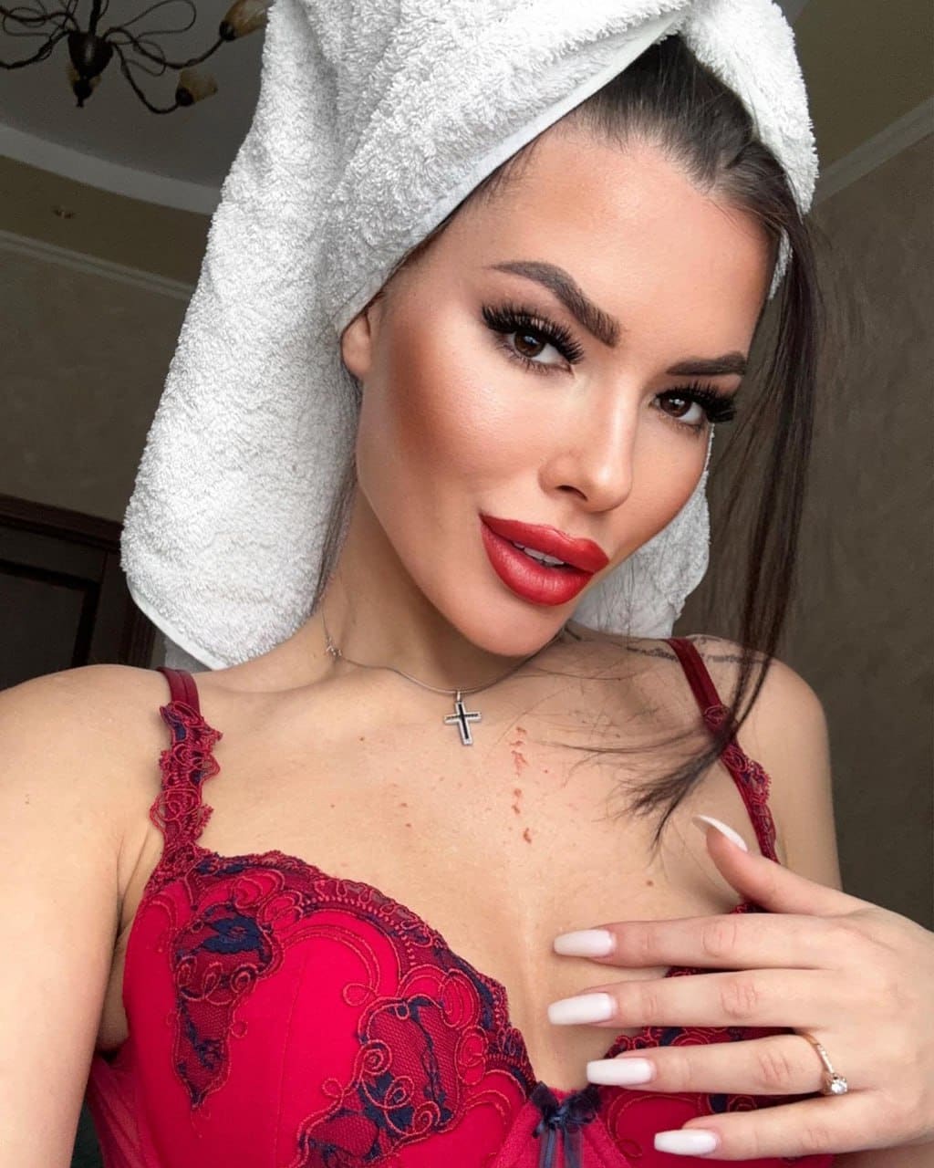 Arina wears a red lace-trimmed lingerie top and cross necklace, wrapped in a white towel, with her hand on her chest indoors.