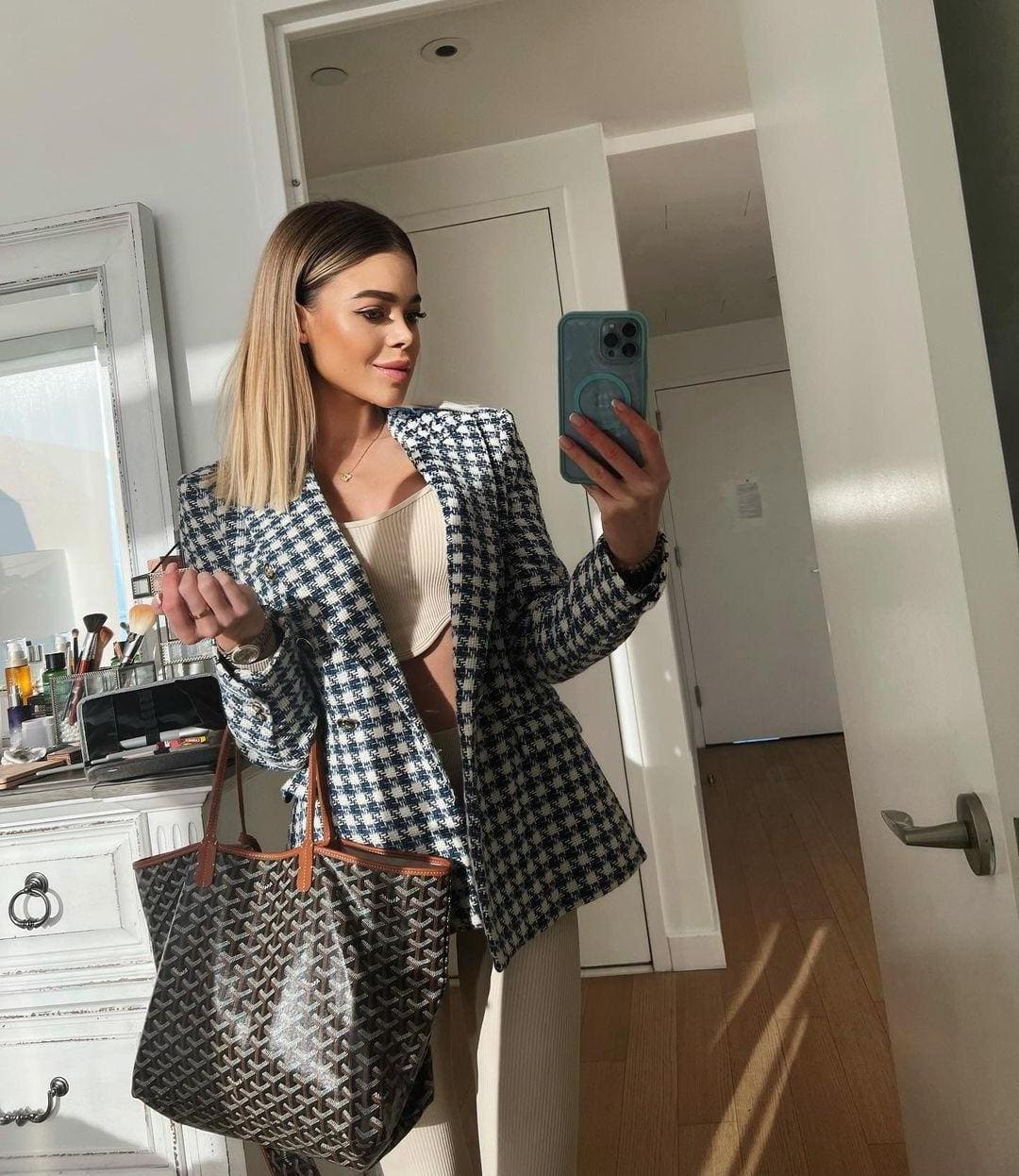 A mirror selfie shows a woman in a houndstooth blazer and beige set, holding a tote bag and phone near a vanity.