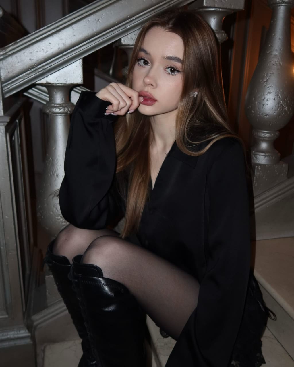 Mariolla sits on an indoor staircase in a black shirt, sheer tights, and knee-high boots, resting a hand near her chin.