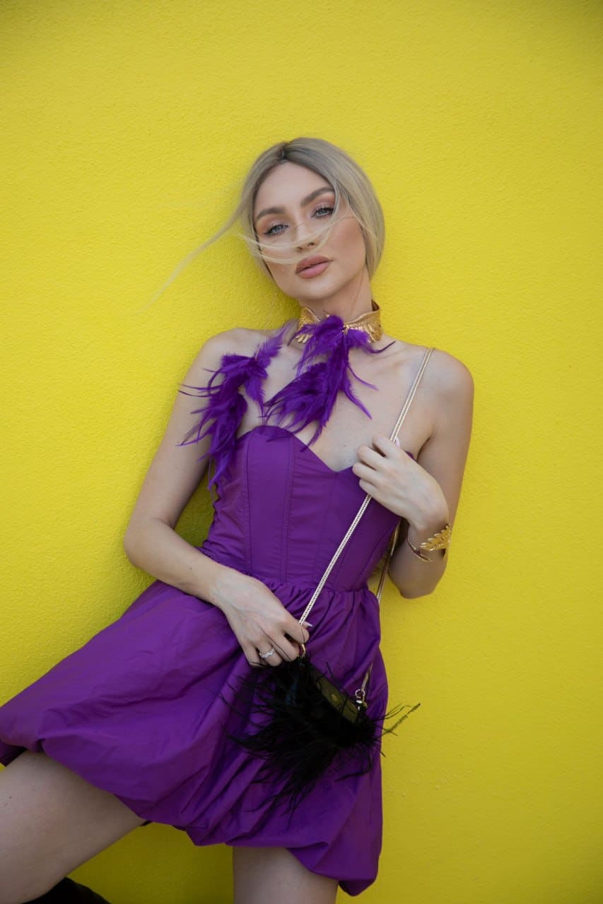 A blonde woman with wind-blown hair poses against a yellow wall in a strapless purple mini dress with a feathered collar, holding a small black handbag.