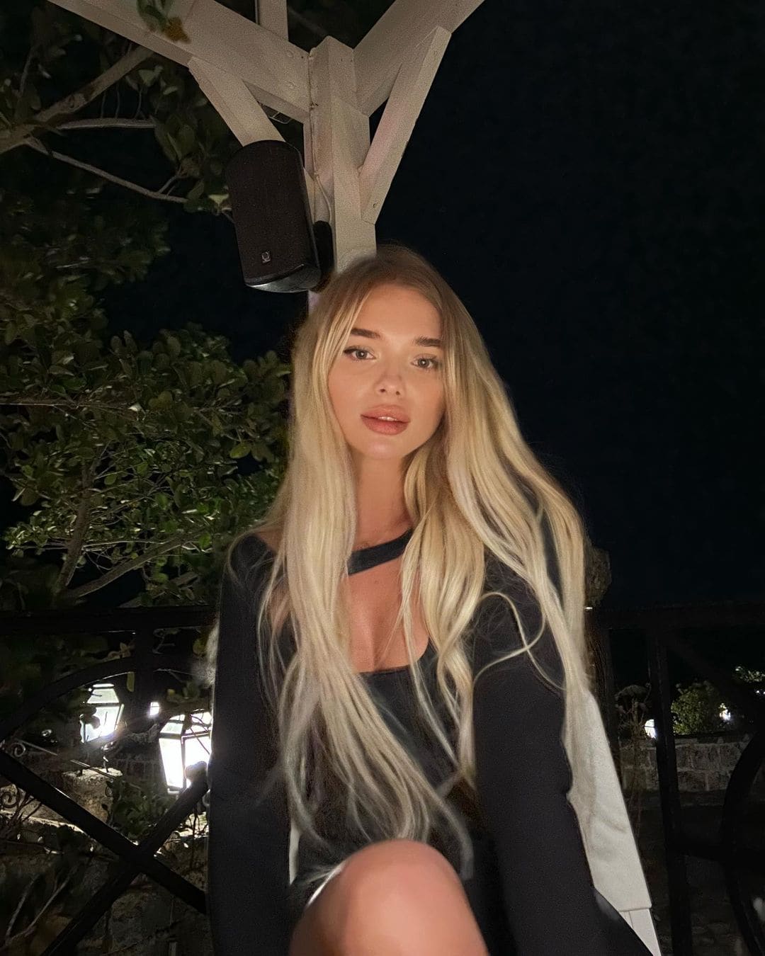 A woman with long wavy blonde hair sits outdoors at night in a black long-sleeved top with a cutout neckline, near a wooden pergola.