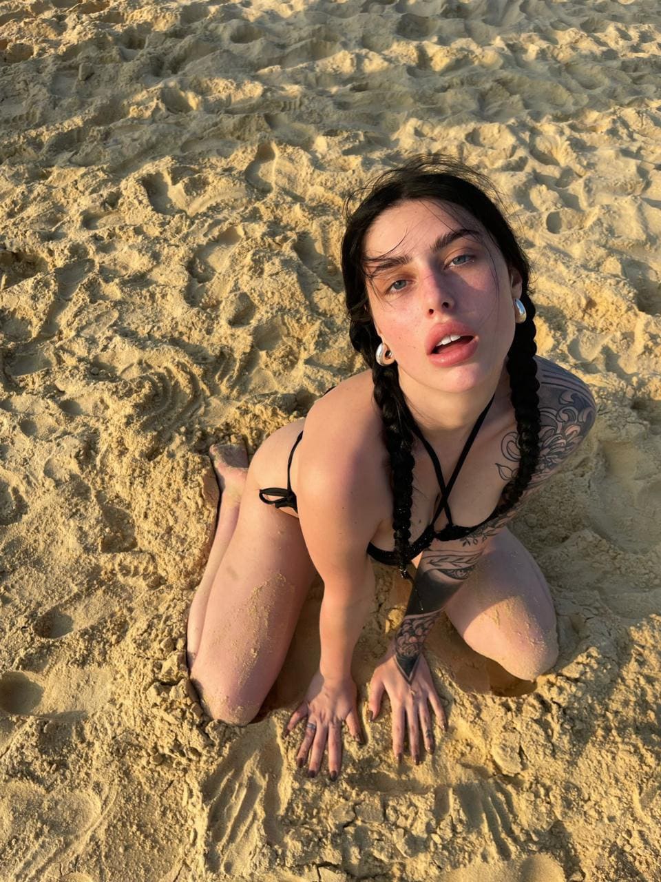 Kneeling on sand, she wears a black bikini top and looks up, with hands pressed into the ground and arm tattoos.