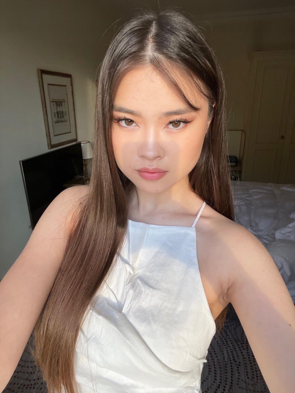 A young woman takes a selfie in a bedroom wearing a white halter top, with a bed and framed picture visible behind her.