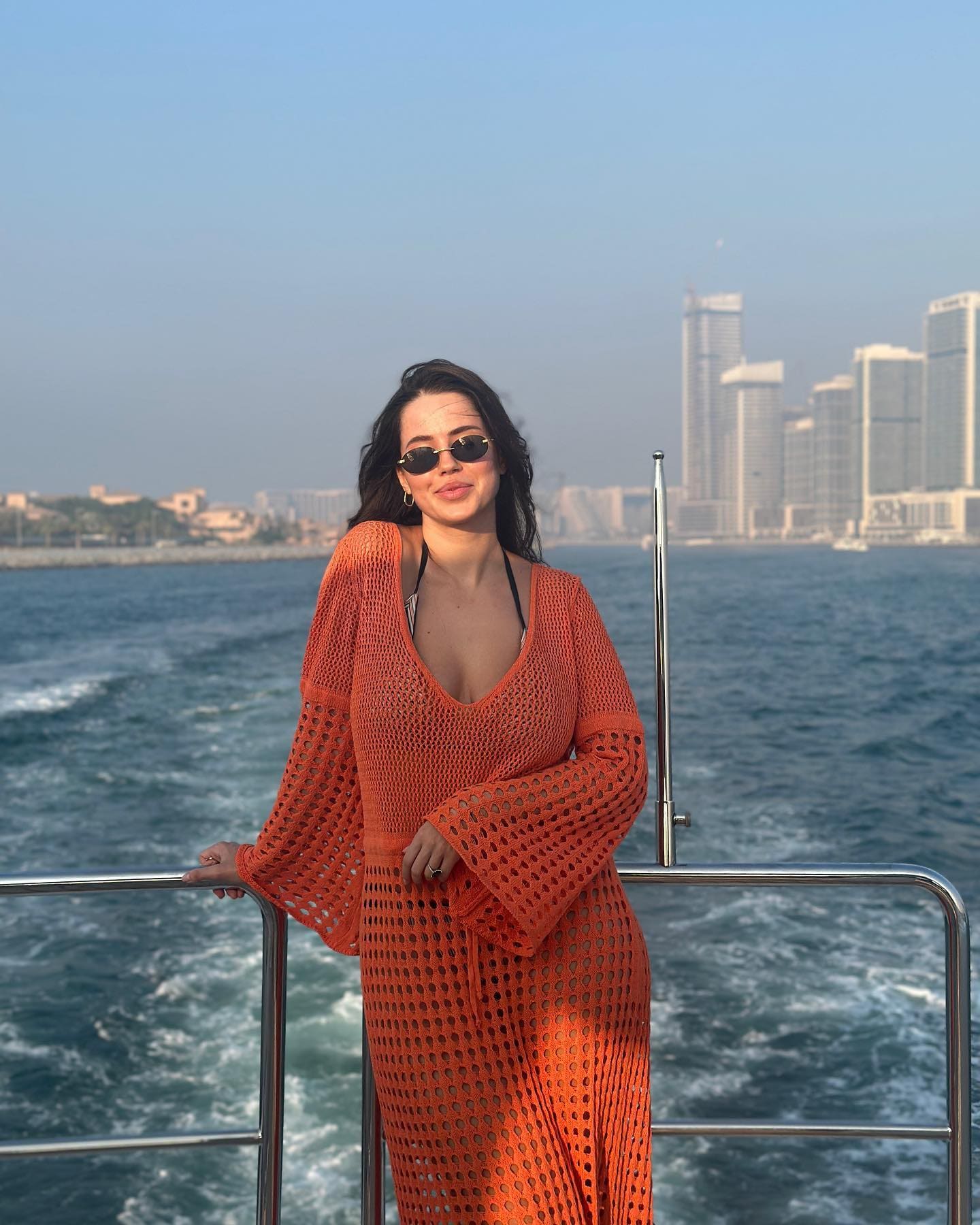 A woman in an orange open-knit dress over a white bikini stands on a boat deck, with sunglasses, water, and a city skyline behind her.