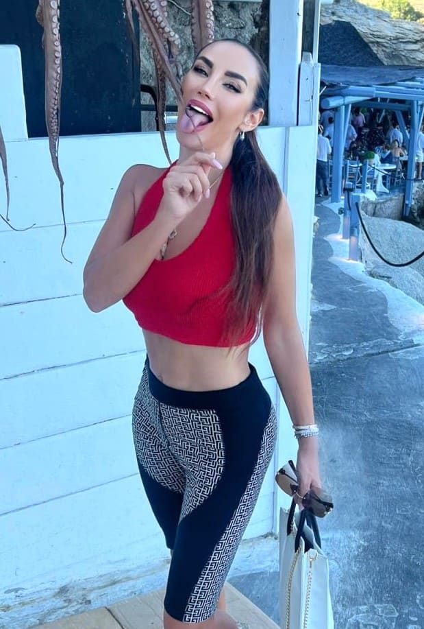 Standing outdoors by a white wall, a woman in a red halter top and patterned leggings holds sunglasses and a bag while sticking her tongue out.