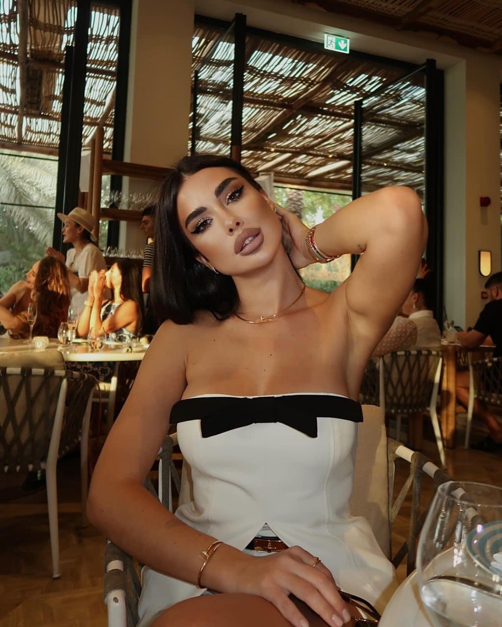 Daniela sits in a restaurant chair wearing a white strapless dress with a black bow, one hand raised to her neck.