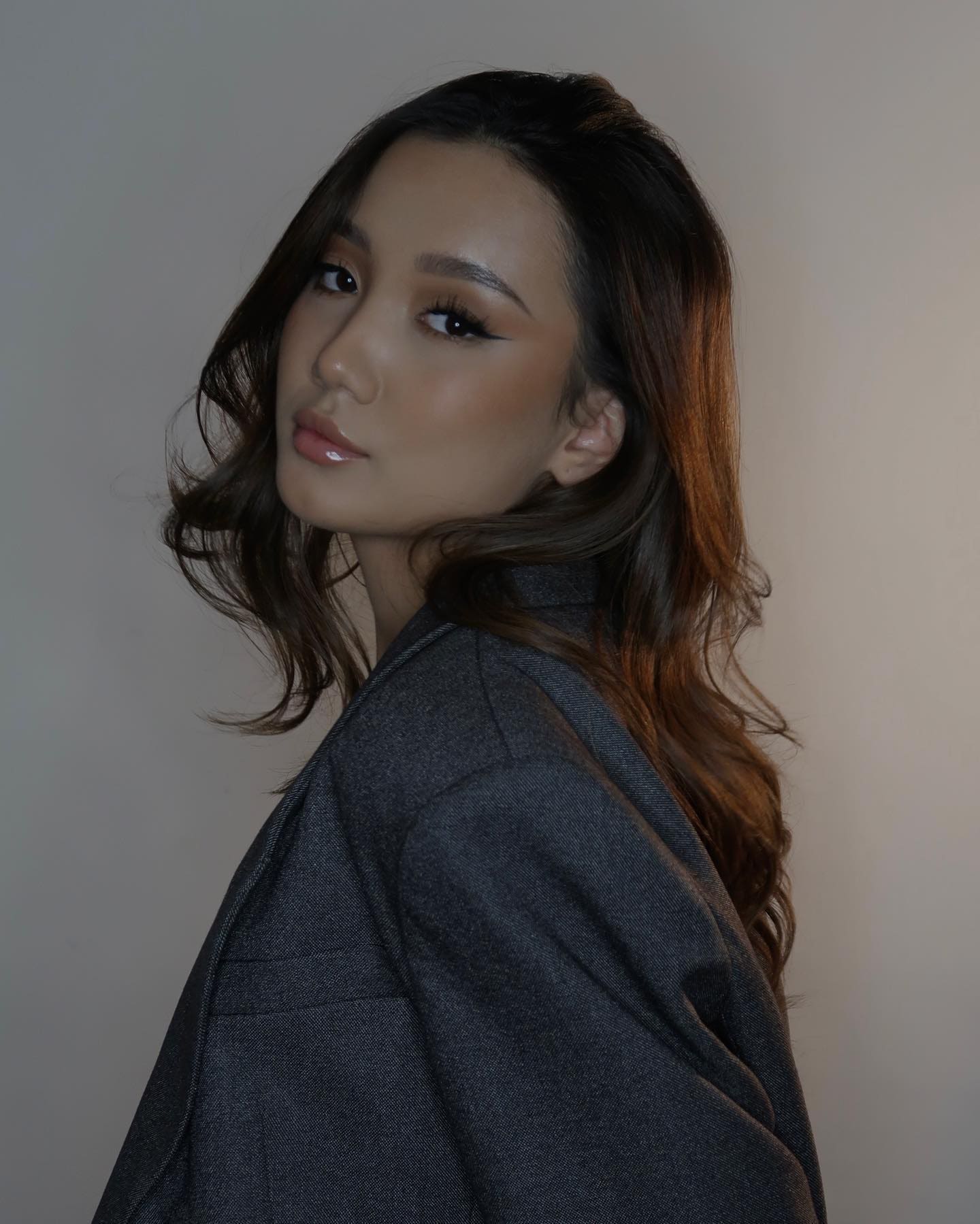 Timina with long dark wavy hair wears a dark grey blazer, looking over her shoulder against a plain light wall.