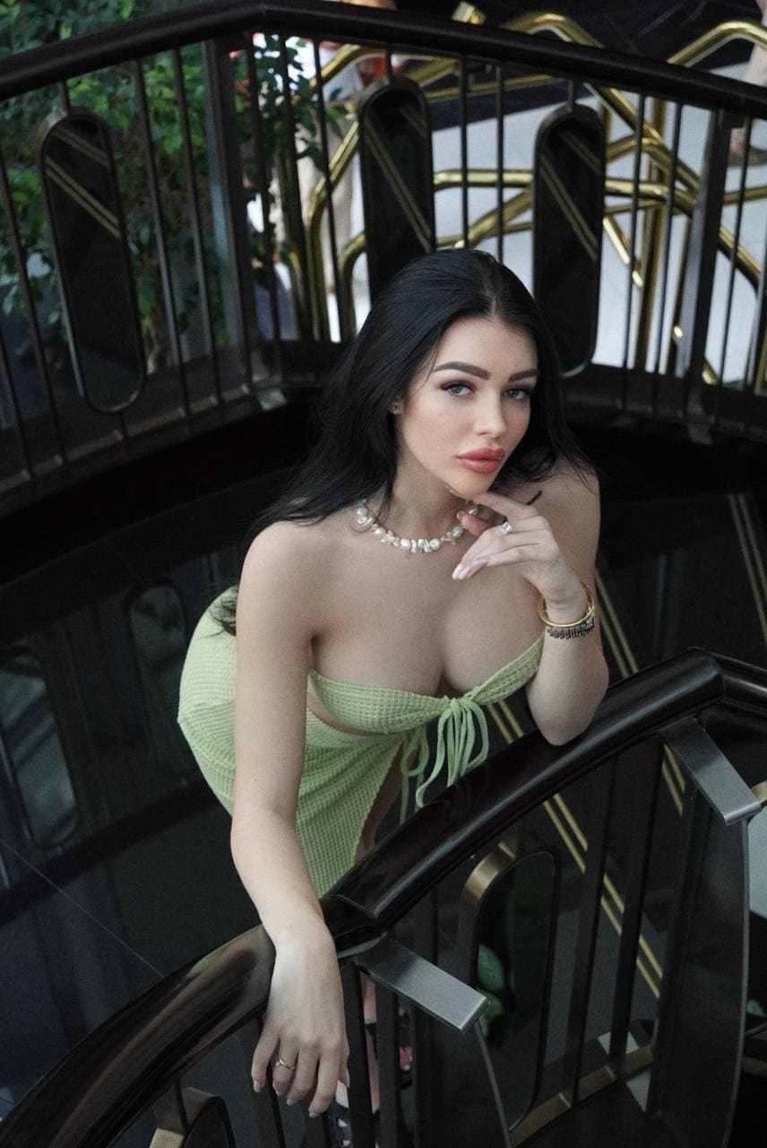 A woman in a green cut-out dress and pearl necklace leans on a black and gold staircase railing, hand near her chin.