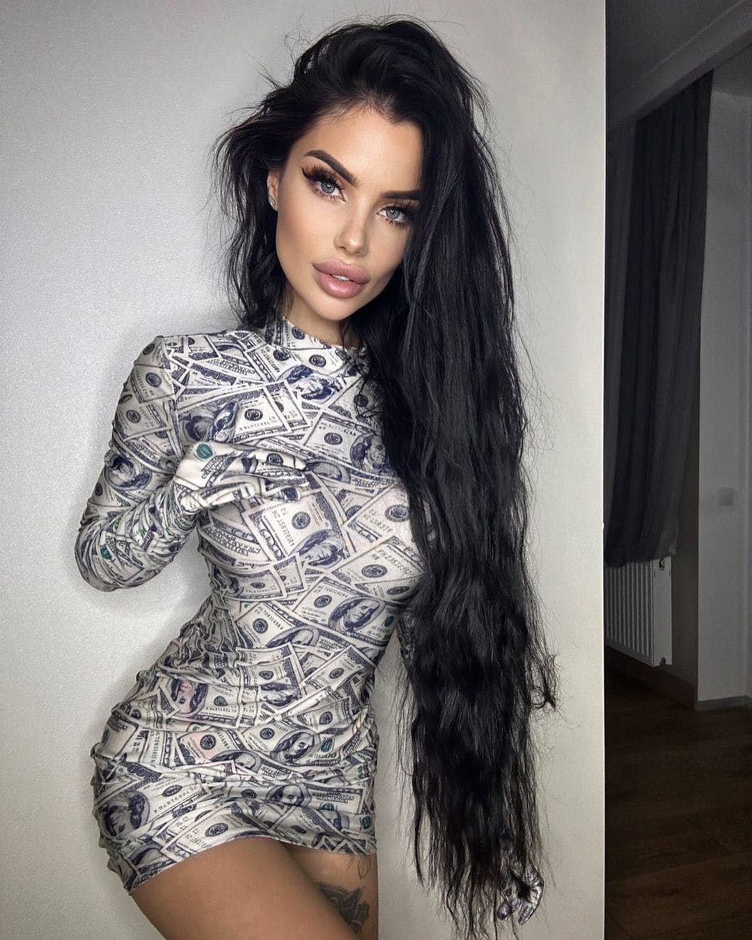 Woman in a banknote-print mini dress against a plain wall, hand near chest, doorway and curtain behind.