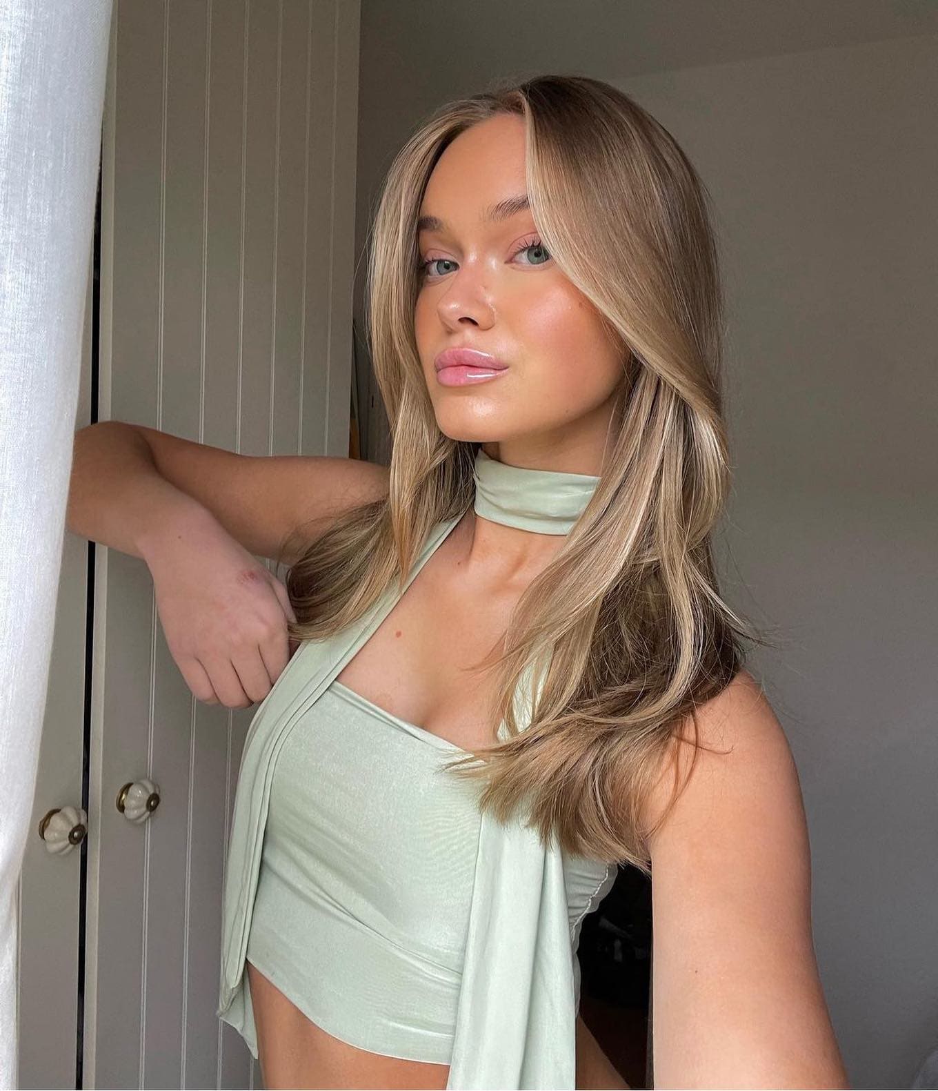 A woman stands indoors next to a white curtain, wearing a light green halter crop top and choker, with her right arm raised and bent.