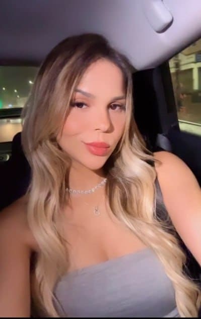 A woman with long blonde hair sits in a car back seat at night, wearing a grey strapless top and looking at the camera.