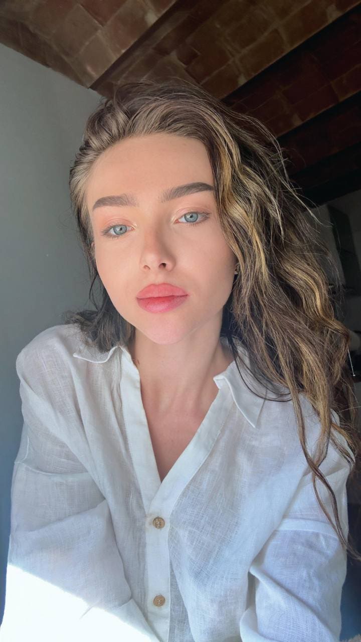 Close-up portrait of a woman in an open white shirt against a brick wall and wooden beams, looking forward.