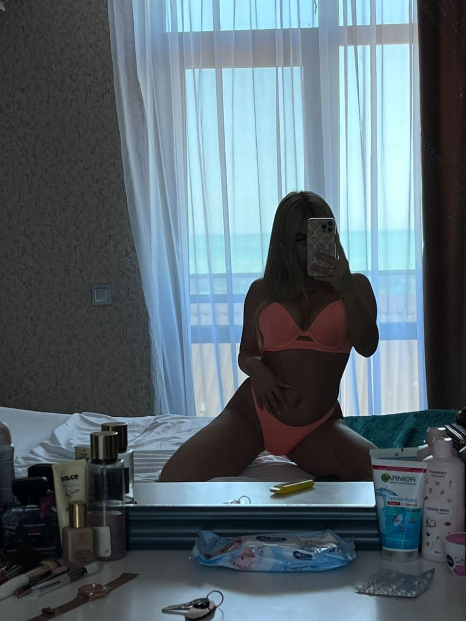 Kneeling on a bed for a mirror selfie, a woman wears a coral bikini, with a vanity of toiletries in the foreground and sheer curtains behind.