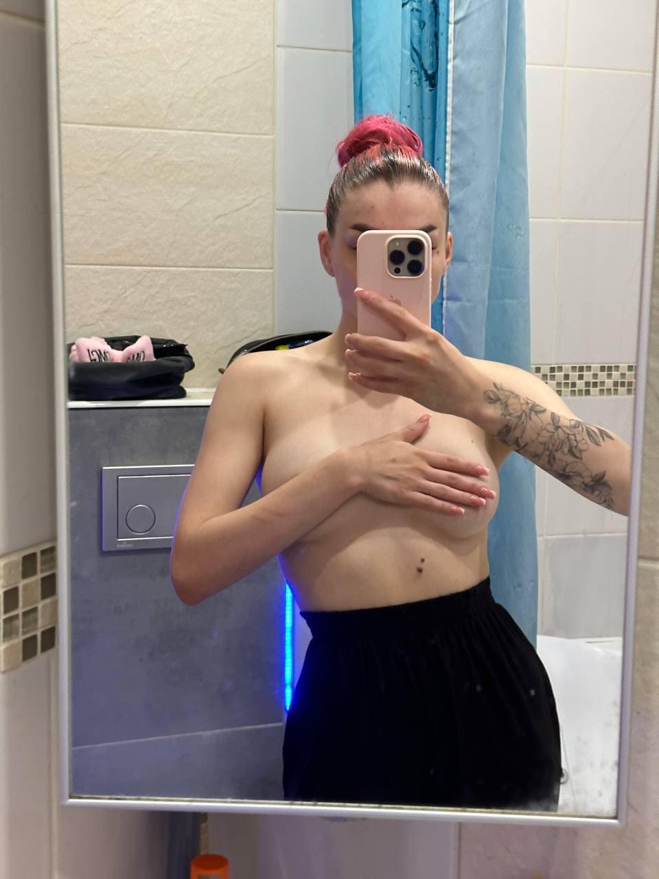 Pink hair in a bun, phone covering face, hand on chest, wearing a black skirt during a bathroom mirror selfie.