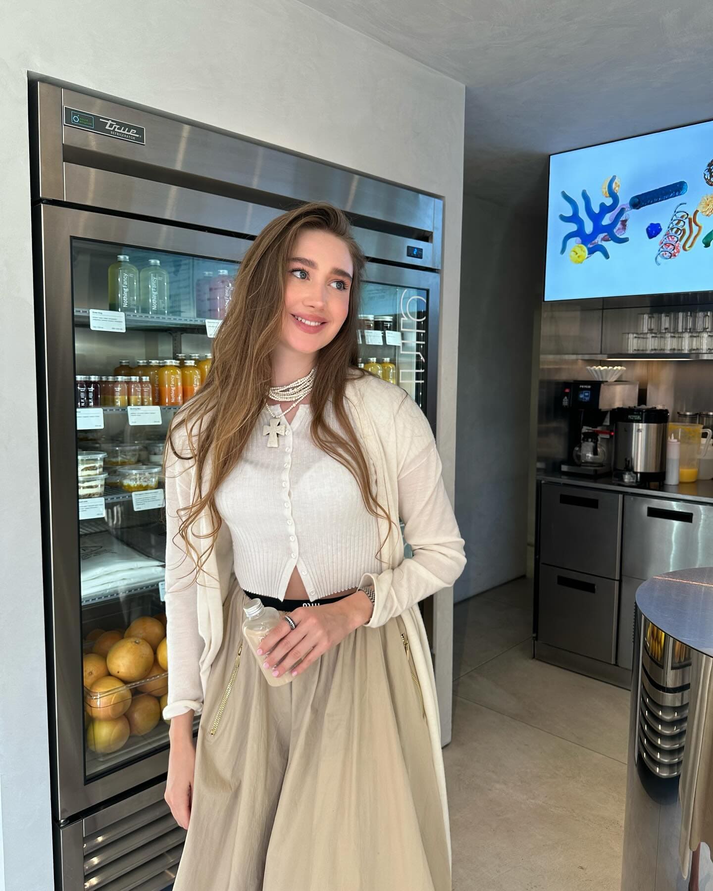 A woman stands in a kitchen holding a small bottle, wearing a white ribbed cardigan, beige skirt, and pearl necklace.