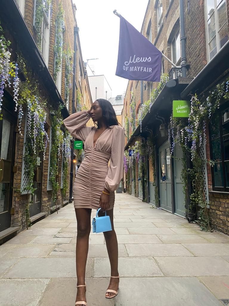 A woman in a beige ruched mini dress and heels stands in a brick alleyway with hanging wisteria.