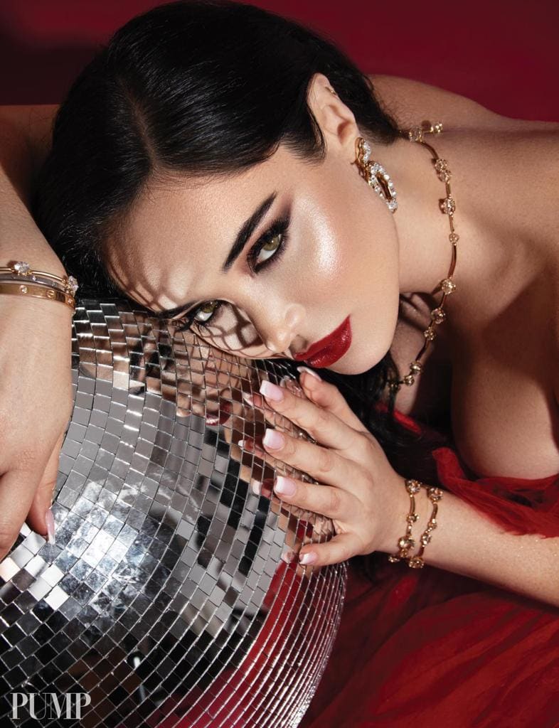 Woman in a red strapless gown and gold jewelry resting her head on a mirrored disco ball against a deep red background.