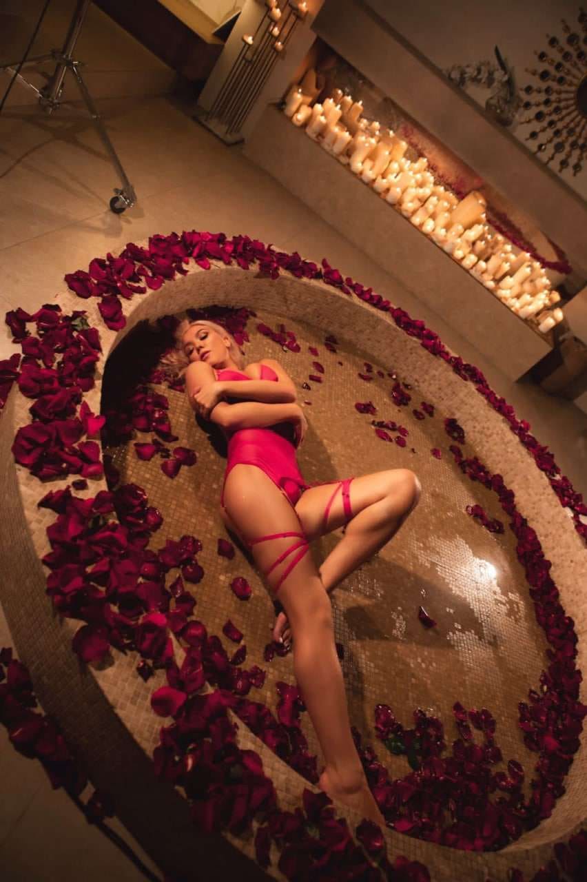 A blonde woman lies in a circular pool with rose petals, wearing a pink swimsuit and garter straps, arms crossed.